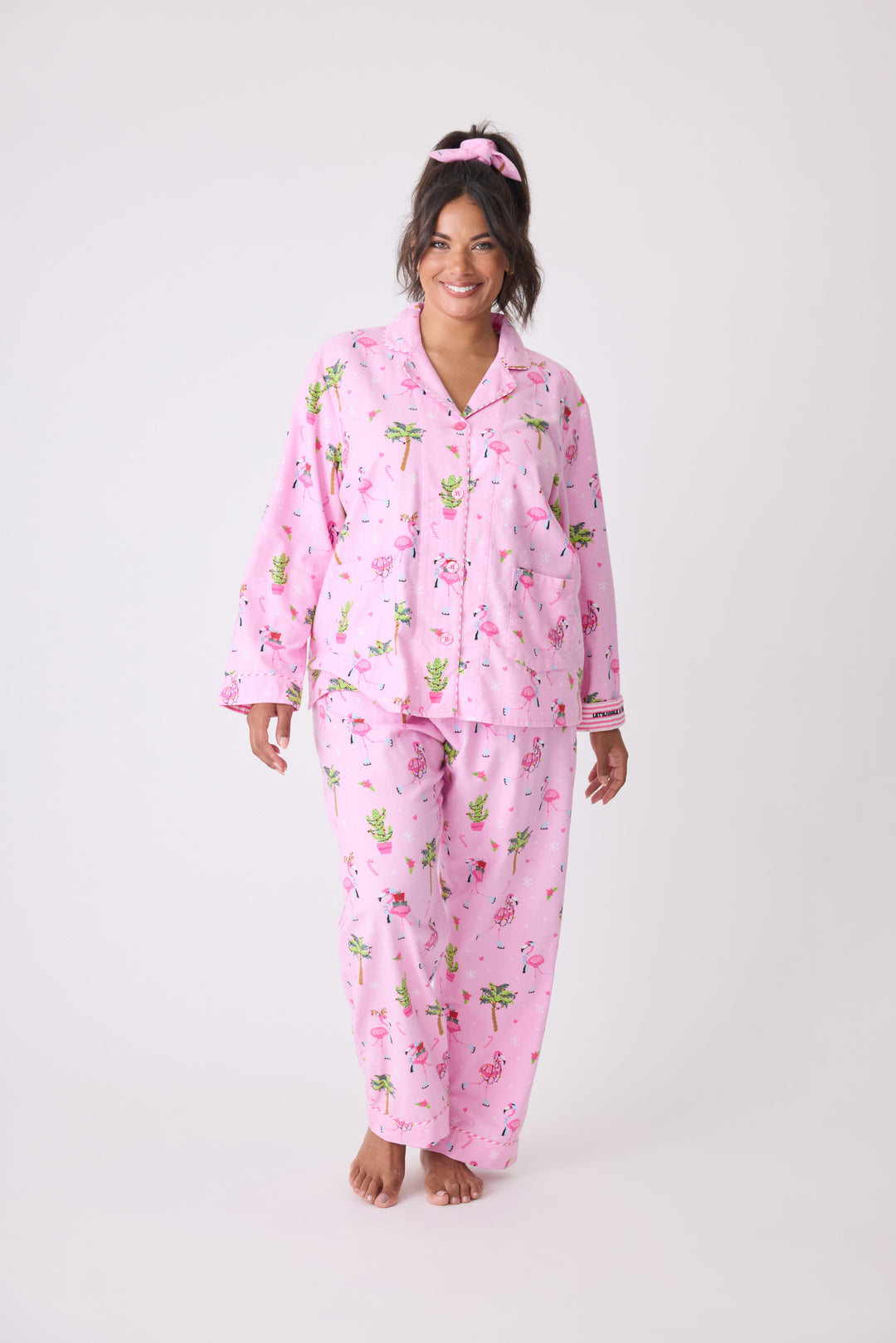 Women's cotton flannel pajama set in pink with retro flamingo print. Button-front collared top & tie-waist pant. Embroidered sleeve cuff "Let's Jingle & Flamingle". Includes matching hair wrap.